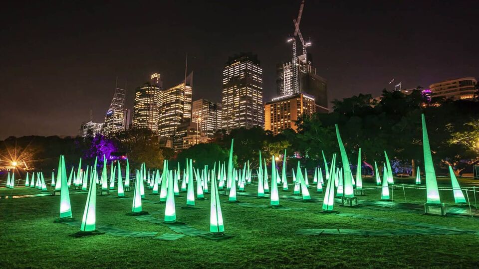 An abundance of green pointy lights in a park, with the city landscpae in the background. Photo taken at night.