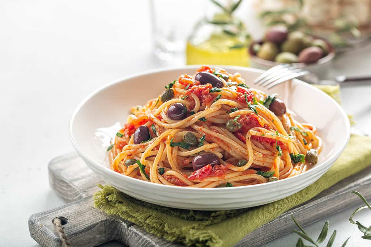 Traditional foods you must try in Italy - Spaghetti alla puttanesca. italian pasta dish with tomatoes, olives, capers and parsley. Light background.
