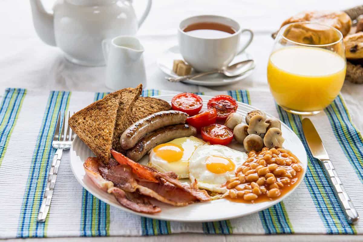 Traditional Full English Breakfast with sunny-side-up fried eggs, bacon, sausages, beans in tomato sauce, toasts, fried tomatoes and mushrooms. Also, with Tea, Orange Juice and Puff Pastry.