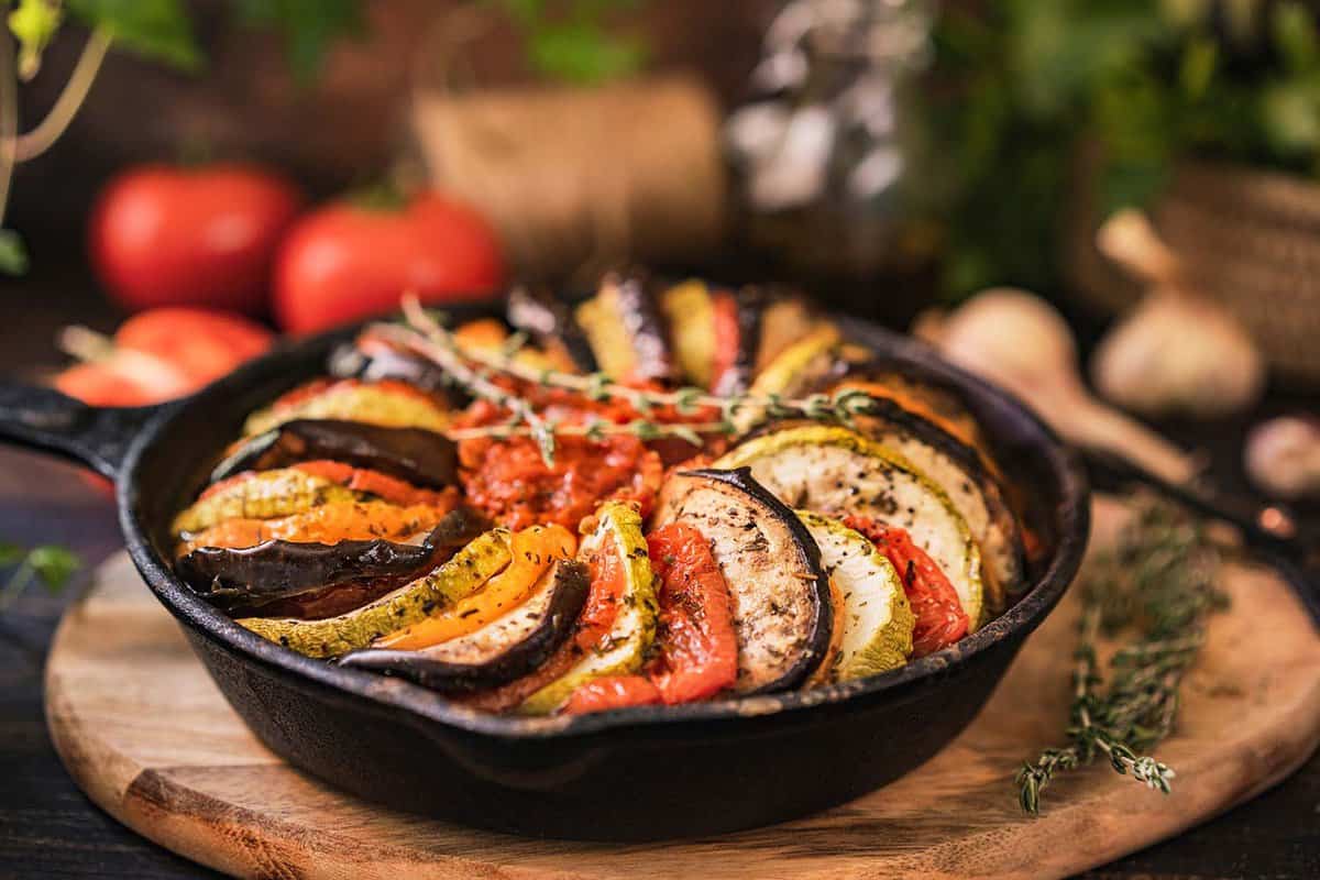 Ratatouille made of zucchini, eggplants, peppers, onions, garlic and tomatoes slices with aromatic herbs. Rustic wooden table.