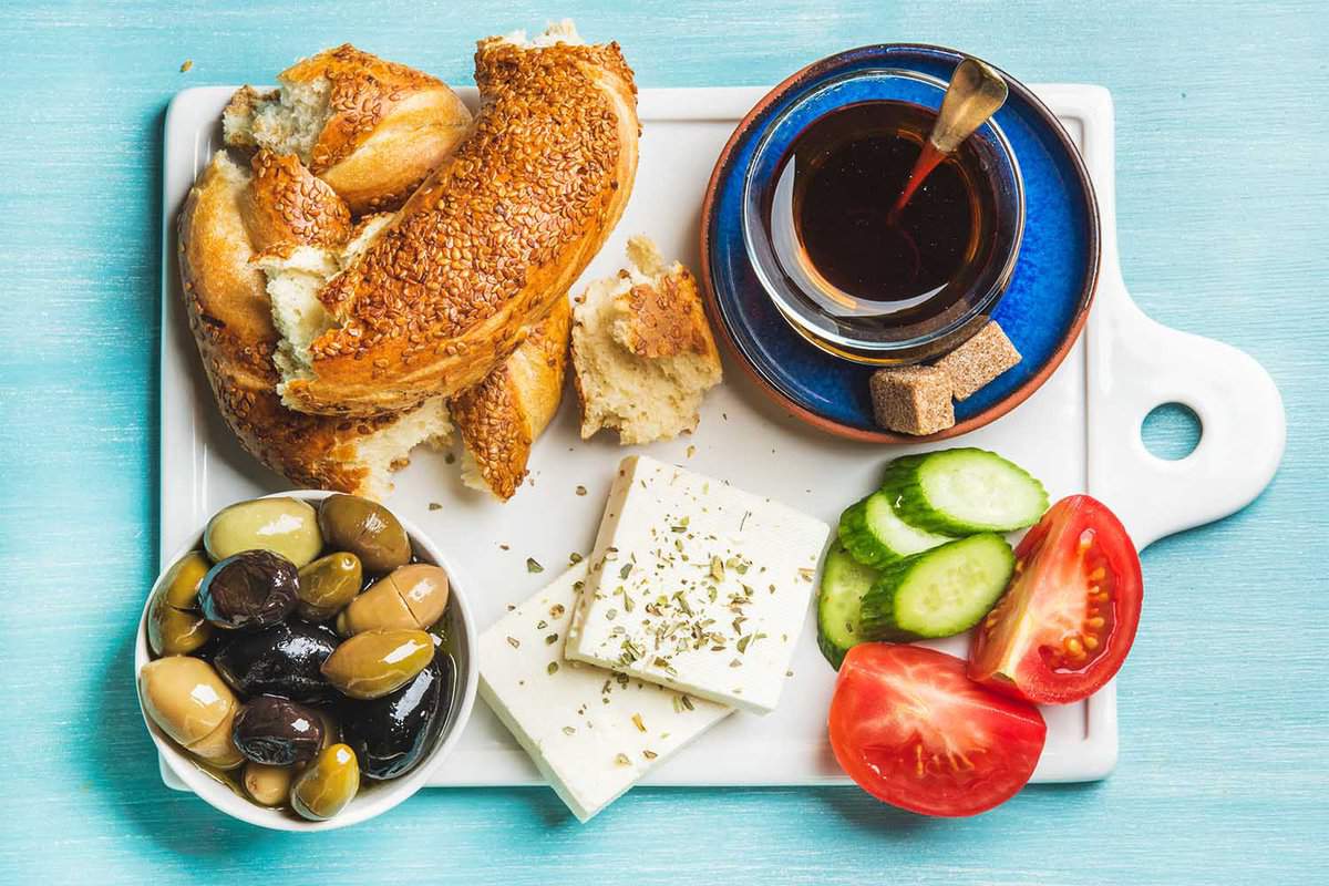 Turkish traditional breakfast with feta cheese, vegetables, olives, simit bagel and tea on white ceramic board over turquoise blue background. Top view