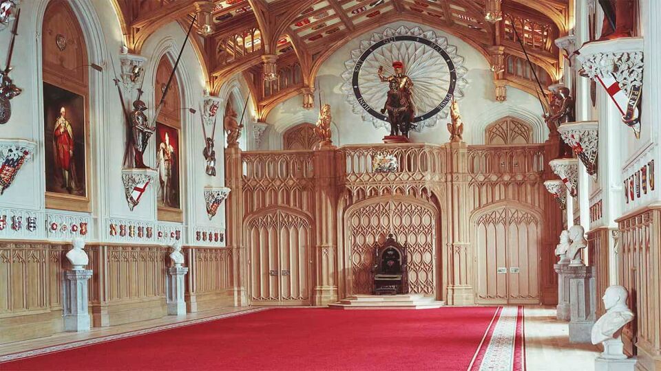 An inside look of St George's Hall in Windsor Castle. There is a contrast between the white, red and golden coloured interior with a red carpet laid in the middle of the room.
