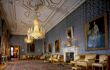 An inside view of the Queen's Ballroom in Windsor Castle. The floorspace has a large patterned carpet in the centre with two golden chandeliers. Alongside the room there are multiple gold coloured chairs with gold framed paintings hung on the walls.