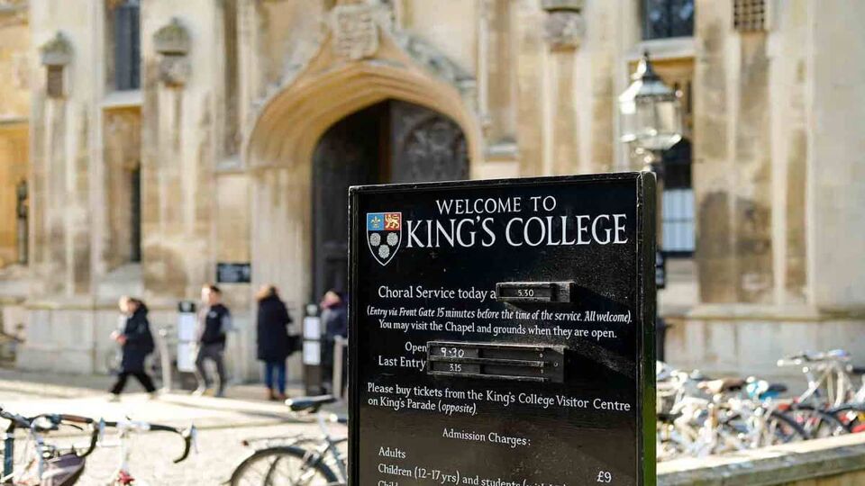 Black sign in front of King's College Cambridge with building and visitors in background