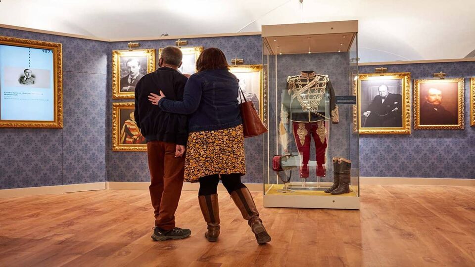 A blue room with lots of paintings and two onlookers staring at a suit of clothes in glass display cabinet