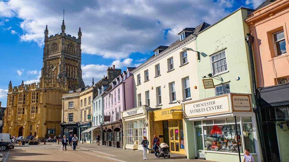 Quaint shops in the centre of Cirencester village, with a large Church in the background, on a sunny day.