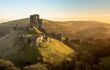 An aerial view of Corfe Castle that sits on a hill during sunset, surrounded large hills in the distance.