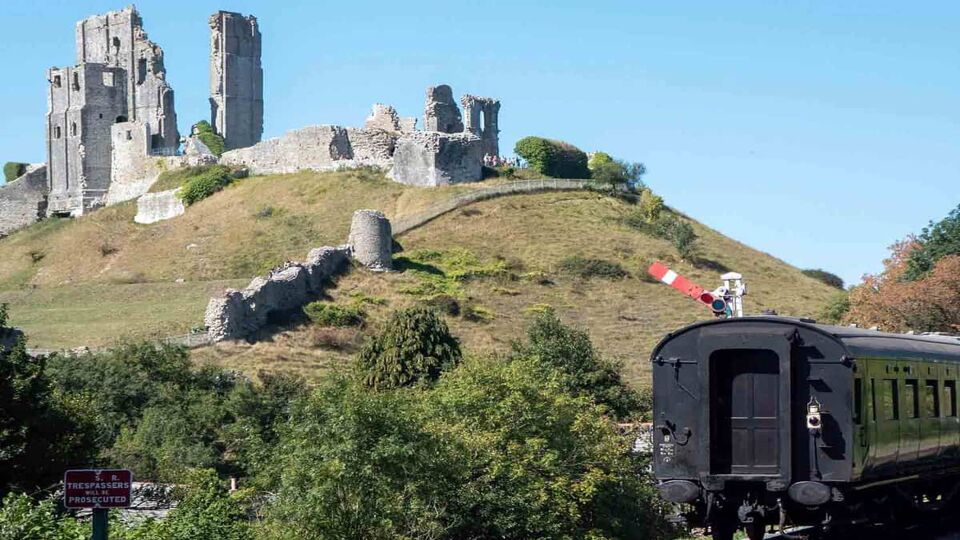 A landscape view of the old castle ruin on the same hill as Corfe Castle but situated below the castle. In the right foreground, there is a train appearing on the railways tracks.