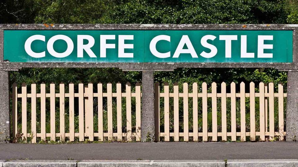 A white Corfe Castle railway station sign on a green background, above a white wooden fence