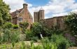 View of the south wing and tower at Sissinghurst Castle Garden, Kent. The tower itself can be seen peeking through the trees in the garden. The garden in the foreground has multiple plants, bushes and trees, with some plants attached on the side of the tower.