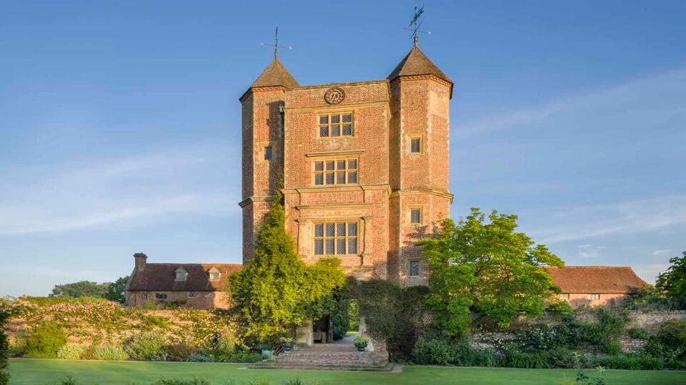 This is an image of the Tower and Lower Courtyard at Sissinghurst Castle Garden in Kent. The Tower has pointed cone shaped roofs and large rectangle windows. The courtyard has well trimmed grass that is seen in the foreground and closer to the entrance of the Tower there are bushes and trees along the sides.