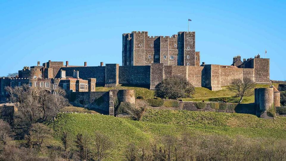 A landscape view to Dover Castle with a blue sky in the background and surrounded by greenery on a raised hill. Dover Castle was founded in the 11th century.