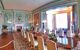 Interior view of a dining room in Leeds Castle. A long polished wood table in the centre with white chairs. The walls are painted in teal with a huge window on the right-hand side, make the room bright.