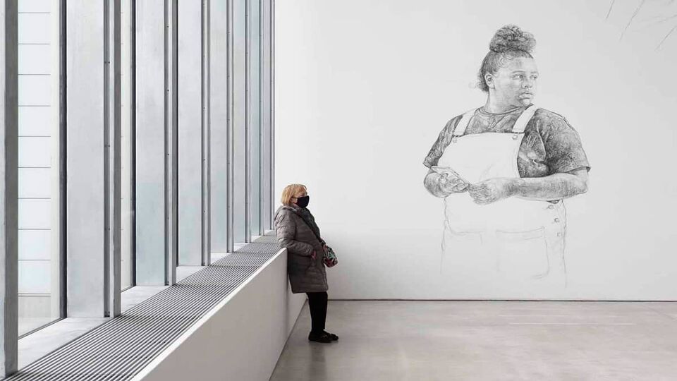 A minimalistic view of inside the gallery where a woman is stook against the side of the wall, next to big glass windows. The room is completely white with an art drawing showcased on the plain white walls.
