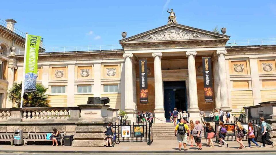 The outside of the Ashmolean Museum. A stately building with pillars on either side of the entrance. There is a crowd of visitors walking up the stairs.