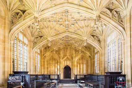 Inside the Bodleian Library Divinity School. The room has dark wooden seating and a vaulted, intricately carved stone ceiling.