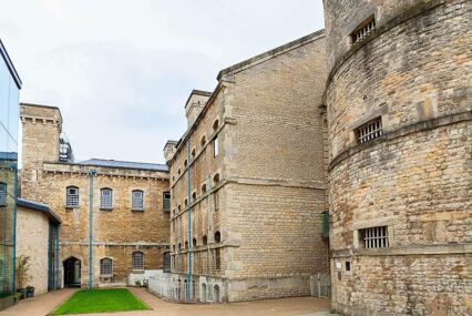 The exterior of Oxford Castle and Prison. Buildings made out of pale stone. The closest building has white painted bars across the windows. There is a small strip of grass in front of the buildings.