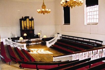 A small room with pale wooden floors, white walls, and two golden chandeliers. Rows of red cushioned tiered seating rise up from a central space. There is a dark wooden organ with golden pipes against the back wall.