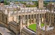 An aerial view of All Souls College cloister. An intricate sandstone building with arched windows and a clockface, has a rectangular grass court in the centre. A public road is visible to the left, and other Oxford buildings and the treeline is visible in the background.