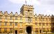 University College gatehouse. An archway sits in the centre, underneath a square turret atop which a flag is flying. The building is long and narrow with triangular peaks in the roof and lines of rectangular windows beneath each small peak. At the bottom of the building, round topiary bushes hang from the walls at regular intervals between the windows.