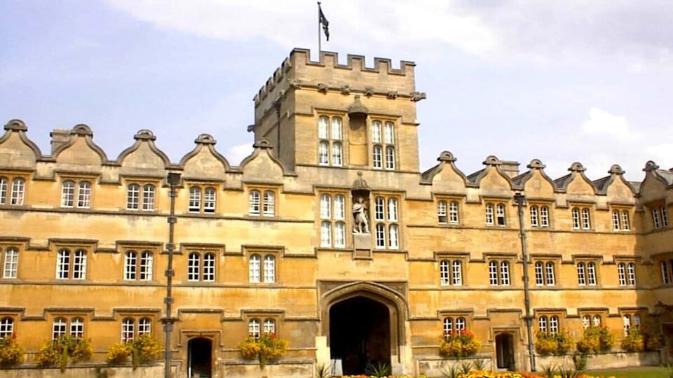 University College gatehouse. An archway sits in the centre, underneath a square turret atop which a flag is flying. The building is long and narrow with triangular peaks in the roof and lines of rectangular windows beneath each small peak. At the bottom of the building, round topiary bushes hang from the walls at regular intervals between the windows.