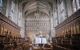 The interior of the Magdalen College chapel. Taken from the plinth, where a open book rests on a bookstand in the shape of a bird. The building has vaulted stone ceilings and arched stained glass windows. In the background, the wall is intricately carved stone in repeating patterns with two figures in the centre towards the ceiling. The lower part of the walls are panelled with pale wood, and two rows of choir pews run along the sides.