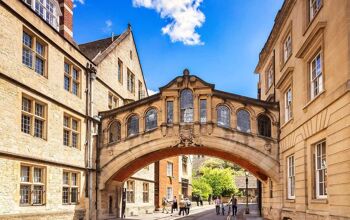 The bridge runs between two buildings above a cobblestone street underneath which people are walking. The bridge is arched sandstone with rectangular glass windows.