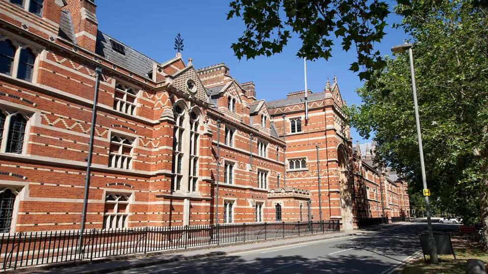 The exterior of a Keble College building. The building consists of red bricks, with white brick interspersed to make a dashed line pattern and a triangular pattern in lines across the building. The building has vertical lines of rectangular windows with white window frames, and a grey slate window. The building backs onto an empty road, with green trees opposite.