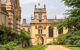 The front quadrangle of Trinity College. The sandstone building in the centre of the image has an archway, tall arched windows, and a small turret with a flag flying in the breeze atop it. The sandstone building to the left has a pattern of faded dark red bricks in stripes along it, and there is neatly trimmed green grass with stripes bordering a path. Green foliage climbs the red-brick patterned building to the left, and a large tree stands on the right of the image.