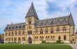 The Oxford University Museum of Natural History building. A narrow yellow sandstone building with many narrow decorative arched windows, a grey slate roof, and a central spire with a grand wooden door. In front of the building is a well-trimmed green lawn and a curved path.