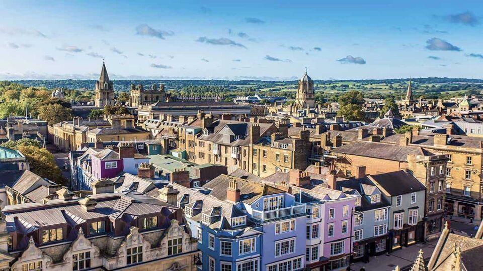 Aerial shot above Oxford. In the foreground there is a row of terraced houses painted in lovely pastel colours. This area of the city is a mix of terraced housing with occasional spires rising up from churches or Oxford University buildings. The majority sandstone buildings are bathed in warm late afternoon sun, and the skyline is a gradient from dark blue to light blue, dotted with small grey clouds.
