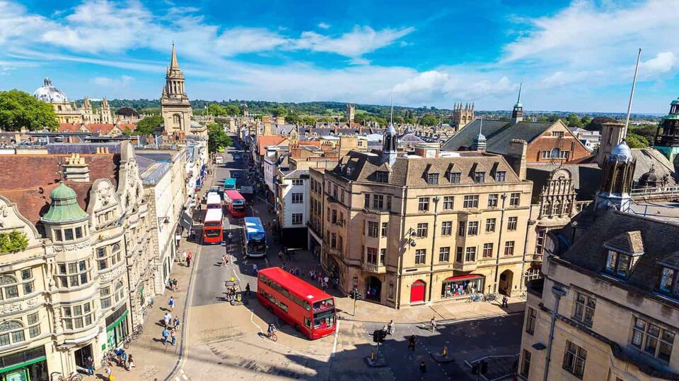 An aerial shot above an intersection in Oxford. A red double-decker bus turns the corner. The buildings are stately and old, most are made of sandstone and have grey or reddish slate roofs. The sky is blue with wispy clouds.