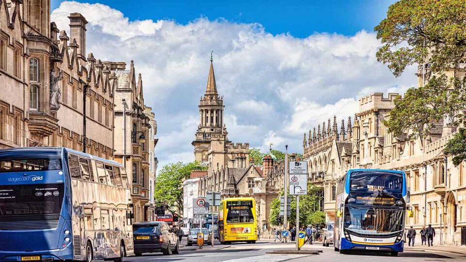 Several blue and yellow double-decker buses go down a wide street in Oxford. The buildings are sandstone, tall, and have intricate details. The sky is blue and there are large fluffy clouds. There are a few trees dotted around, with green leaves beginning to turn orange.
