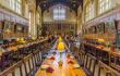 Inside the great hall of Christ Church college - the inspiration for Hogwarts dining hall. The arched ceilings have dark wooden beams with windows beneath them. The walls underneath the windows are panelled with wood and have oil portraits hanging. There are three long wooden tables with wooden chairs along them down the length of the hall. Each table has lamps along the centre with yellow lampshades, and crockery and cutlery laid out ready for dining. Next to each plate is a red serviette folded to stand up prettily.