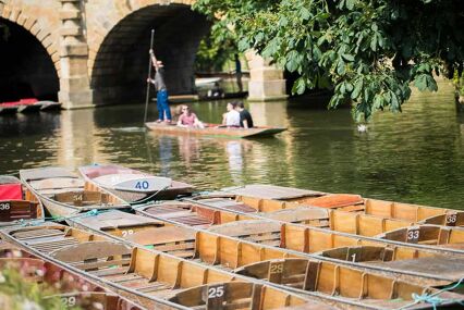 Four numbered wooden punts (boats) lined up next to each other on the river's edge. In the background there is a bridge with two arches, and a man standing, pushing a punt along with a large pole. There are two passengers in his punt.