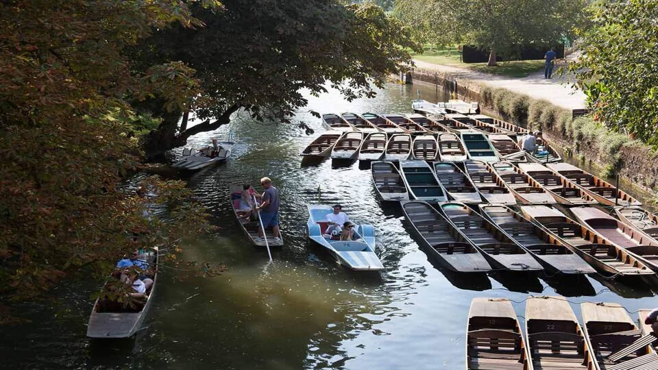 Many punting boats are lined up on the right side of the river. There is a path next to the river, where a person wearing blue clothing is standing. On the left side of the river, four punts with passengers are moving along. The area is shaded by green, low-hanging trees.