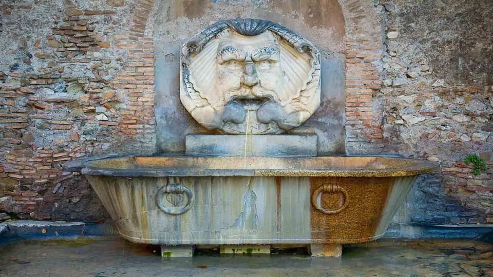 roman fountain with a strange fat face on the wall above an old bath tub