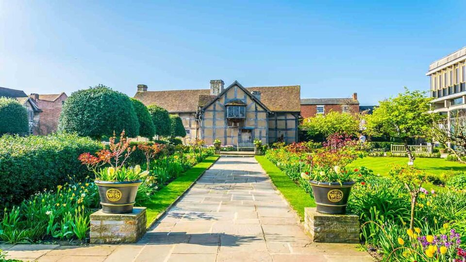 A front view of the birth house of William Shakespeare on a sunny day in Stratford upon Avon, England. A large bricked pathway leading to the entrance of the house with beautifully arranged flowers and plants alongside this walkway.