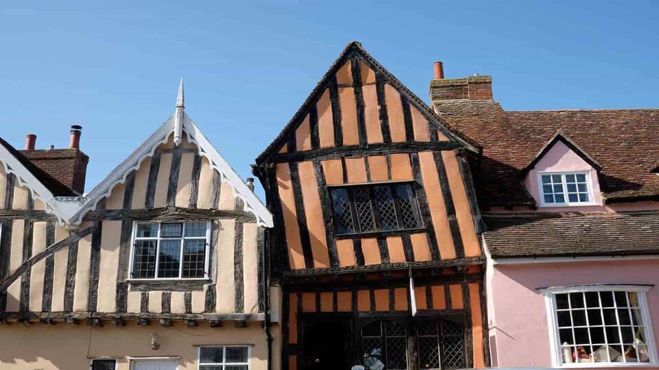 A low angle view of three medieval houses. From left to right, the first one is painted yellow, the second is painted orange and the third is in pink. The photo highlights the triangle shaped roofings that can be seen on all houses, which is quite distinct to the Middle Ages. .