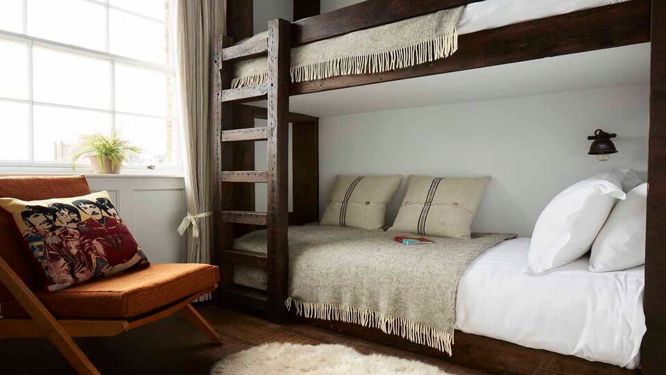 A bunk bed made of dark wood, made up with white sheets and a beige blanket and pillows. A cushioned chair sits next to the bunk beds, and a small plant sits on the windowsill.