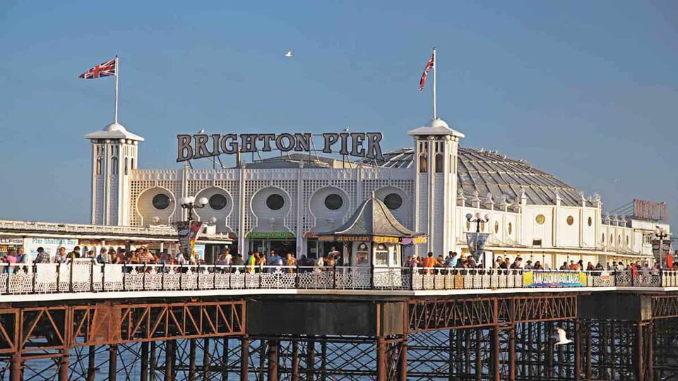 The entrance to Brighton pier. There is a sign saying 'Brighton Pier' above the pier building (which is painted white, and has two pillars bearing flags on the left and right of the building). You can see visitors walking around on the pier, and the pier supports going into the sea.
