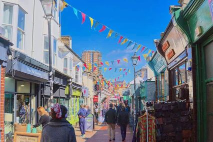 A vibrant shopping street full of people on a clear day. Colourful bunting hangs from building to building.