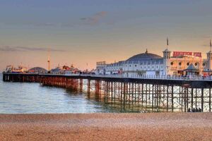 Brighton Pier at sunset. Fairground rides and entrance to the pier visible above the reflective water.
