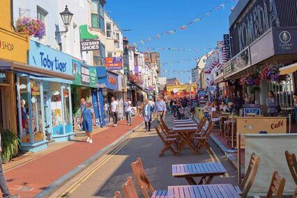 The shops in Brighton Lanes. Taken on a sunny day, the eclectic shops have brightly painted shopfront signs, and there is colourful bunting criss-crossing between the building roofs. The sky is blue, and several cafes have outdoor seating in the pedestrianised street. Shoppers walk on the pavement in front of the shops.