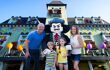 A family of four smiling for a photo with a Lego mascot against a green house with blue skies