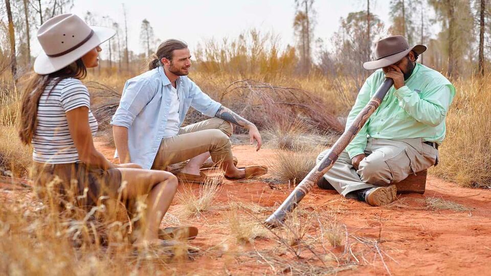 A man blowing into a A man playing the didgeridoo and a couple listening, in the desert.