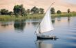 Felucca sail boat sailing across the smooth Nile River toward a verdant green shore with desert background in Egypt