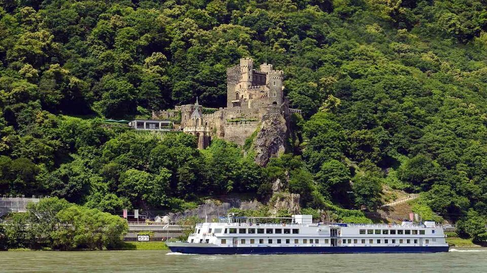small runiend castle on a forested hill, small river cruise ship sailing past