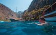 Man swimming next to traditional Turkish Gulets in the blue Mediterranean water. The cliffs on either side form butterfly valley in the background.