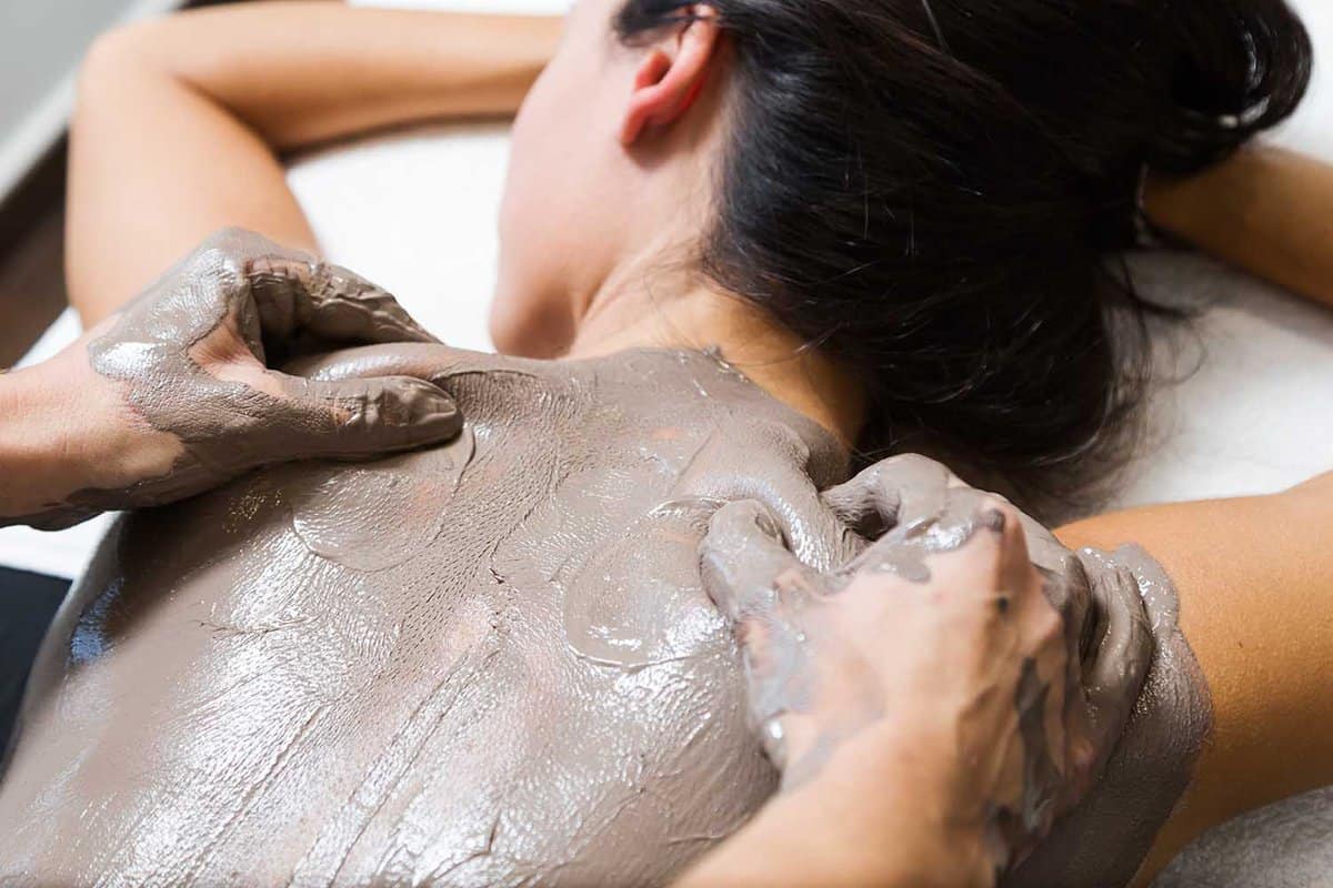 Portrait of beautiful young woman having clay body mask apply by beautician. Detox ritual.